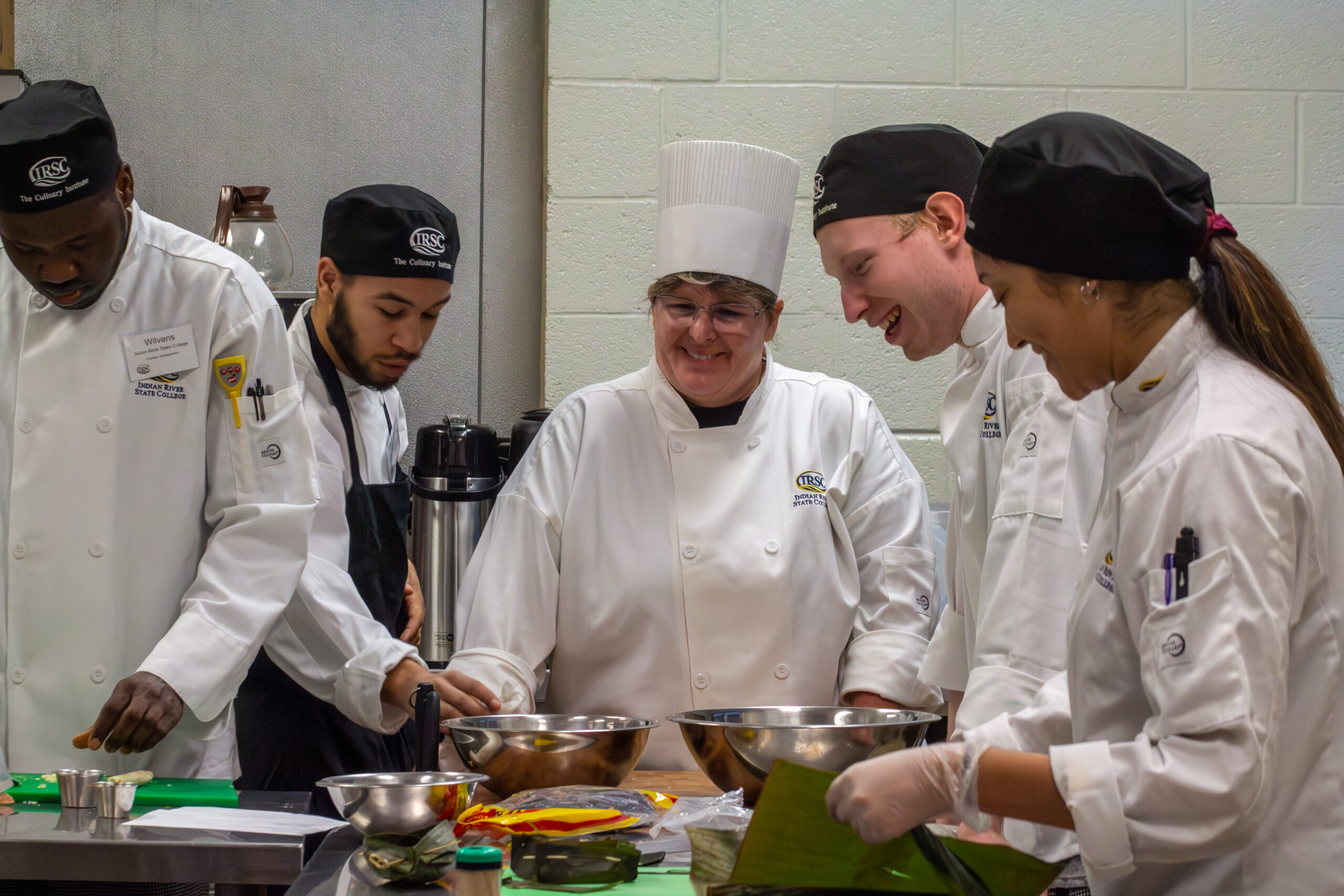 A photo showing a cuisine professor and four college students in a kitchen. The photo shows them gathering around a prep table with food.