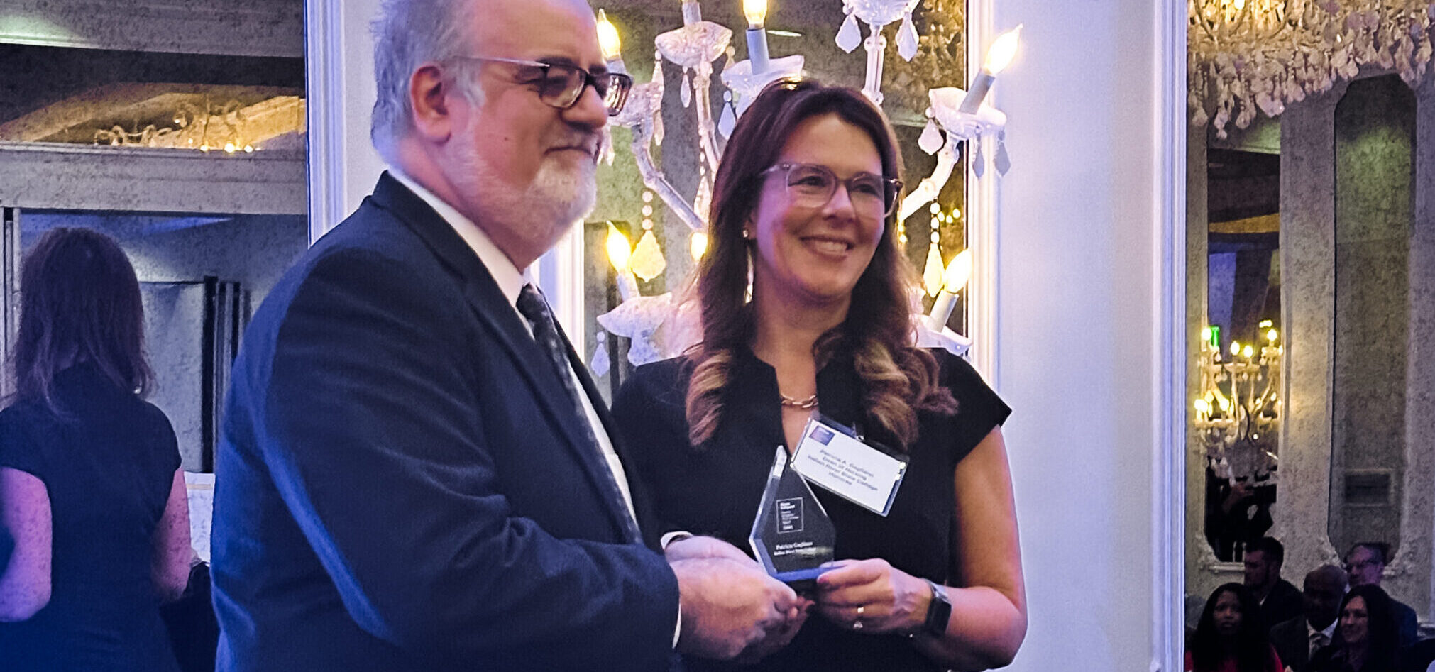 A woman in a black dress smiles while receiving an award from a man in a suit at a formal event. They stand on stage under elegant lighting, with rows of awards visible on the table behind them.